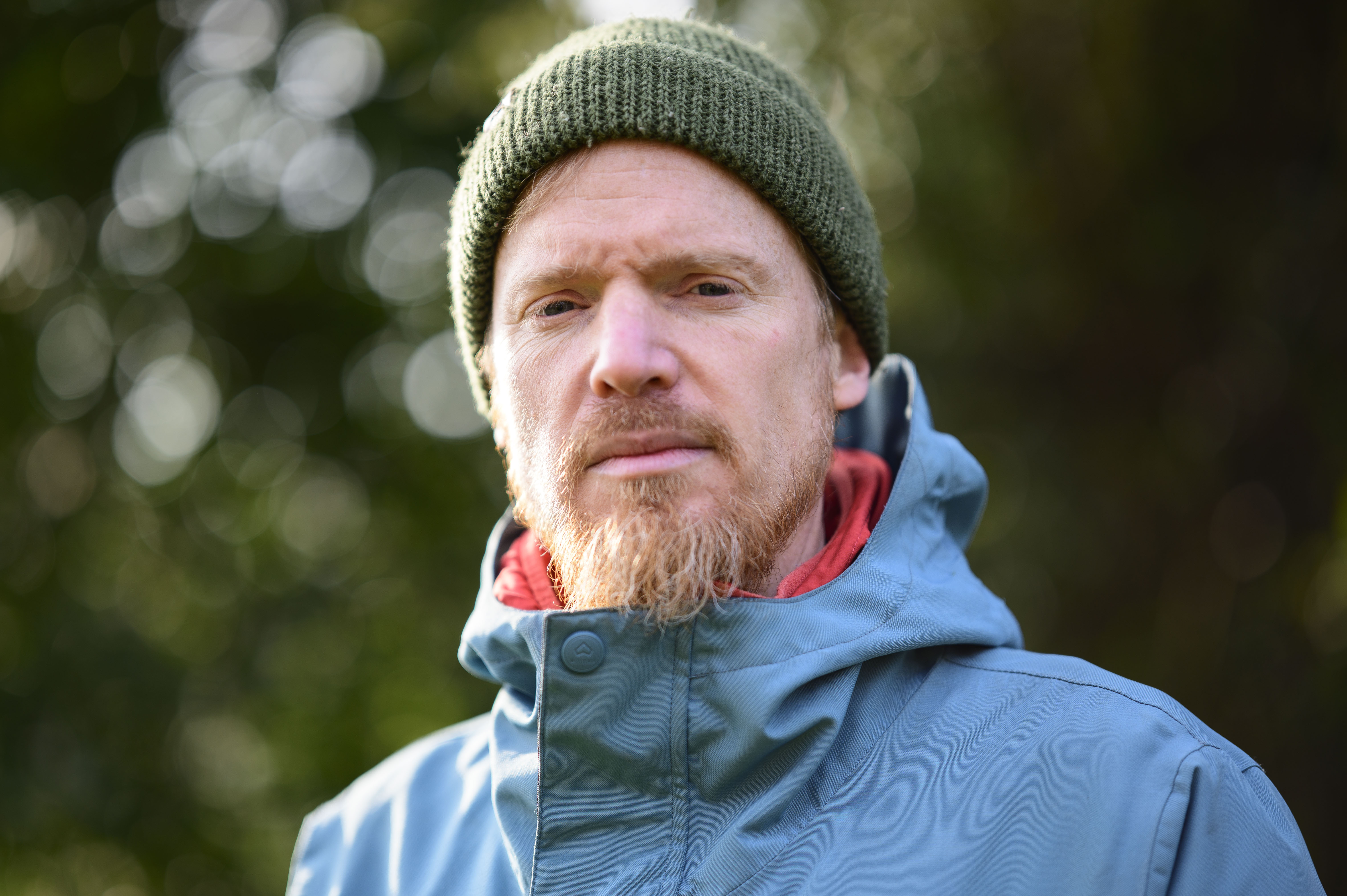 Portrait of a man in a blue coat and green beanie, with dappled light through a background tree