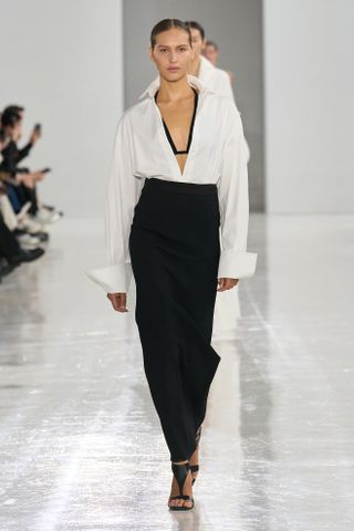 A photo from the Max Mara S/S 25 runway show of a model wearing a white button-down shirt with a black bikini underneath and a black high-waist maxi skirt.