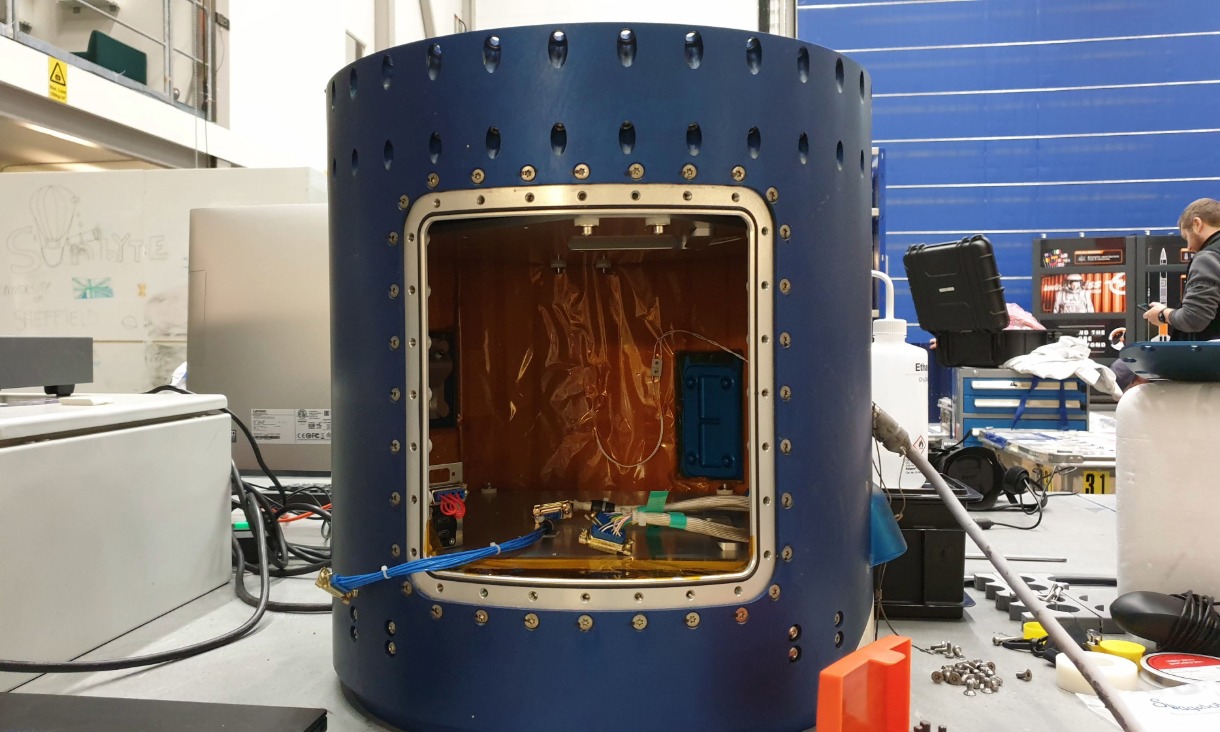 a blue cylinder with a glass window sits in a hangar surrounded by wires and metal boxes