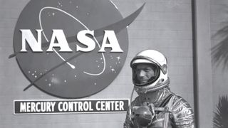 Black and white image of a man in a shiny space suit stands to the right, facing left, in front of a brick wall with a large NASA meagball logo hung, with a sign beneath that reads, 'mercury control center'