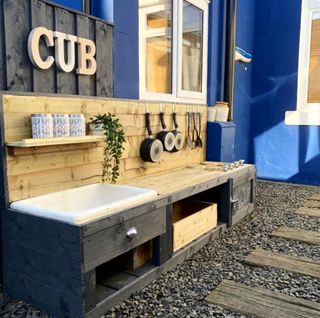 Mud kitchen made from fence panels and painted silver-grey, complete with shelf, plant and pots and pans