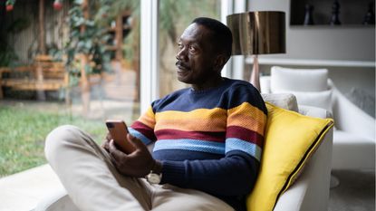 A pensive-looking older man sits next to a window and looks outside while also holding a smartphone.