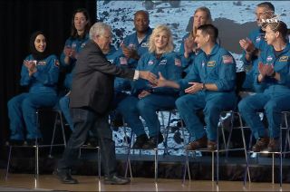 a man in a dark suit shakes hands with a sitting man in a blue flight suit while other people in blue flight suits smile