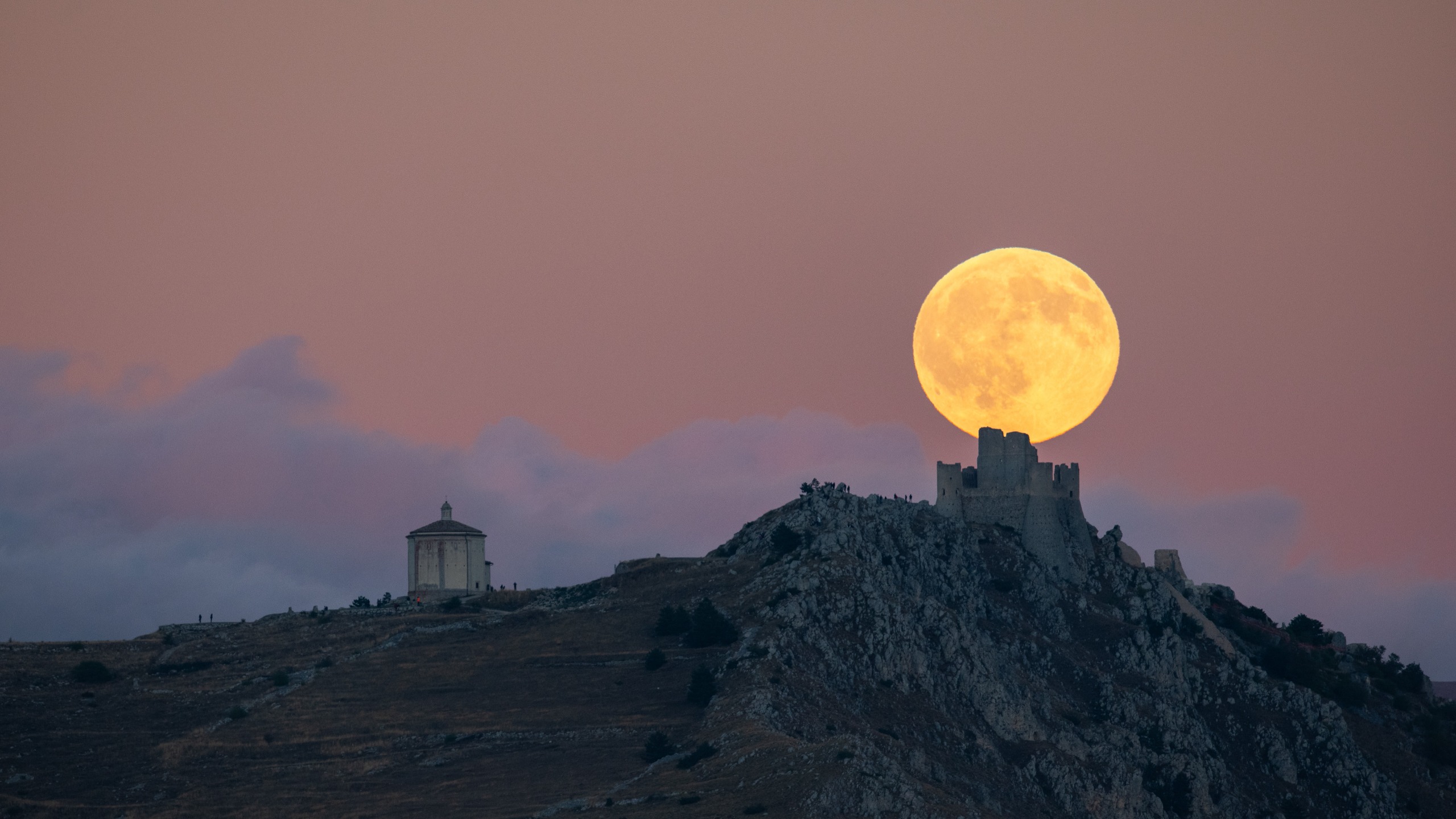A large golden full moon rises behind the silhouette of Rocca Calascio Castle and the Church of Santa Maria della Piet&amp;agrave; in Calascio, Italy.