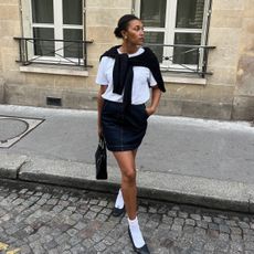 Influencer Lena Farl wears a denim mini skirt and a white crewneck with white socks and black ballet flats. 