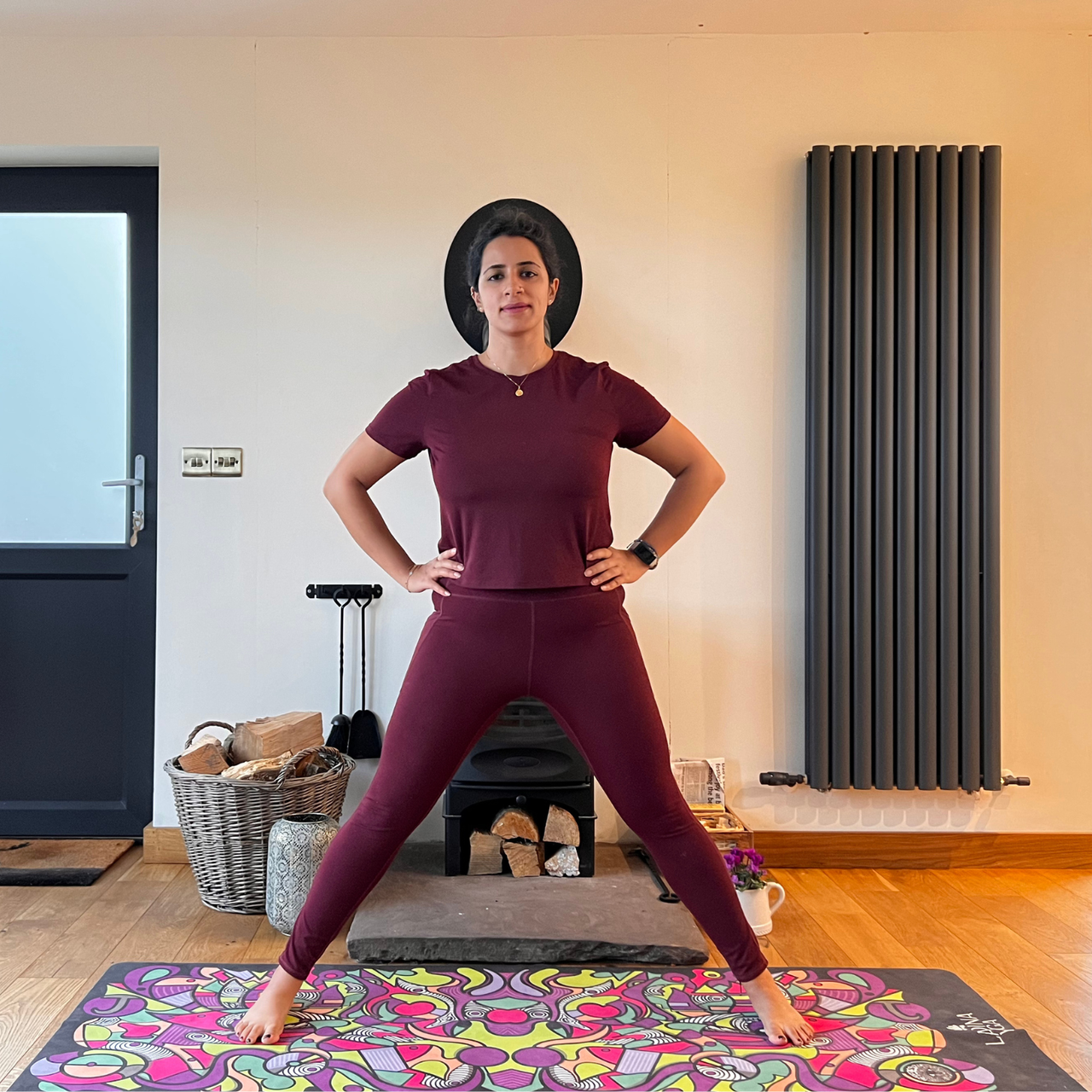 Maysun Hassanaly performs a wide legged forward fold. She is standing on a colorful yoga mat with her legs wide apart and hands on hip.