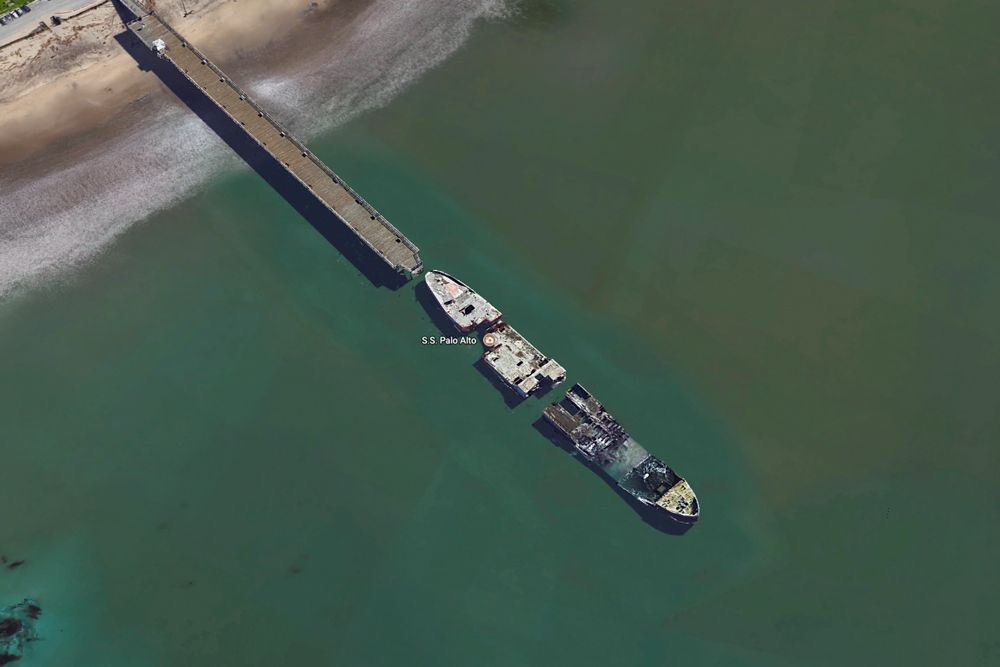 We see a bird&#039;s-eye view of a long broke military ship floating at the end of a dock on a beach.