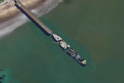 We see a bird's-eye view of a long broke military ship floating at the end of a dock on a beach.
