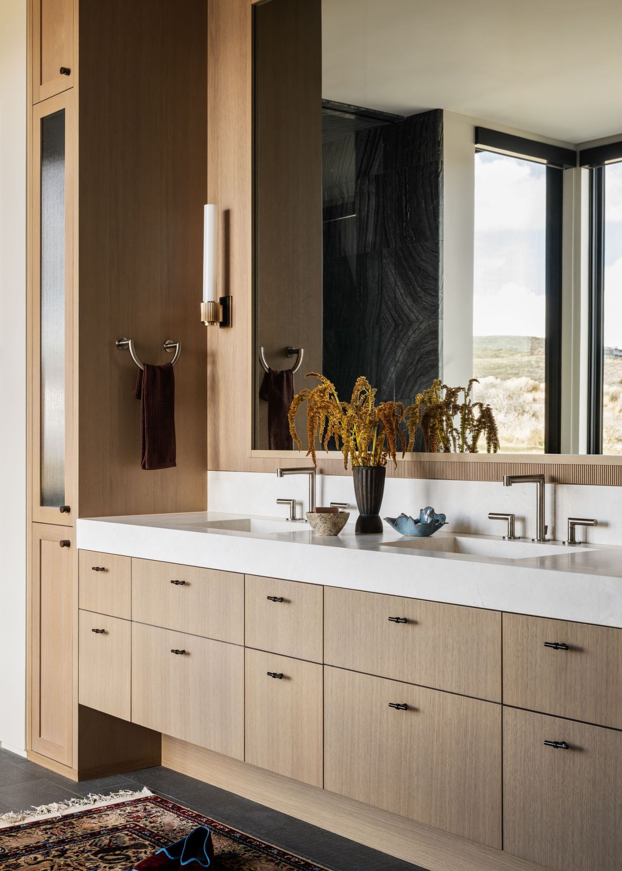 A modern bathroom with light wood built in cabinetry and a large mirror above a double vanity unit and an antique rug in front