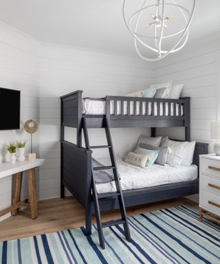 Sophisticated bunk bed set-up in modern coastal bedroom, with white horizontal wall paneling, white caged orb light, and wall mounted TV.