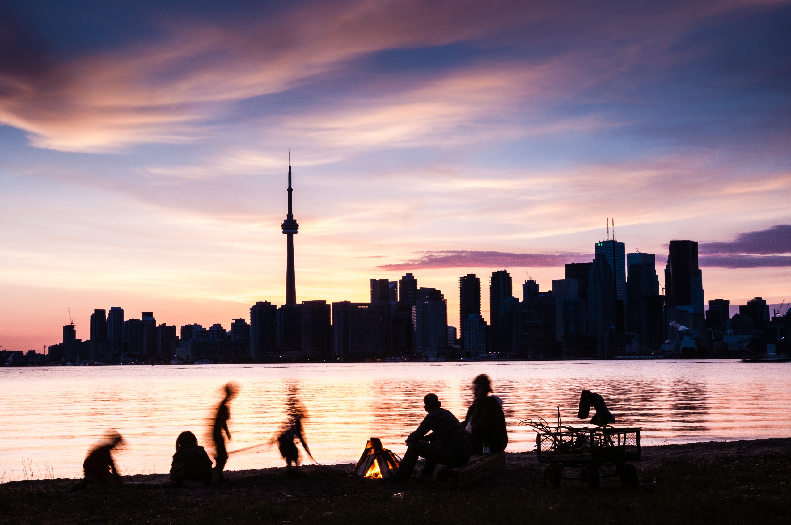 A pink sunset view capturing a skyline as a black silhouette in front of which a series of people lies on the beach with a fire.