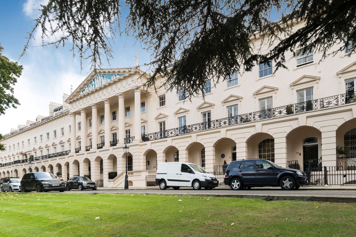 A large Georgian/Regent style white home with a street of cars in front and large balconies and pillars stand on the front of the home