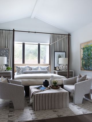 master bedroom with canopy bed in front of windows, two accent chairs and striped coffee table