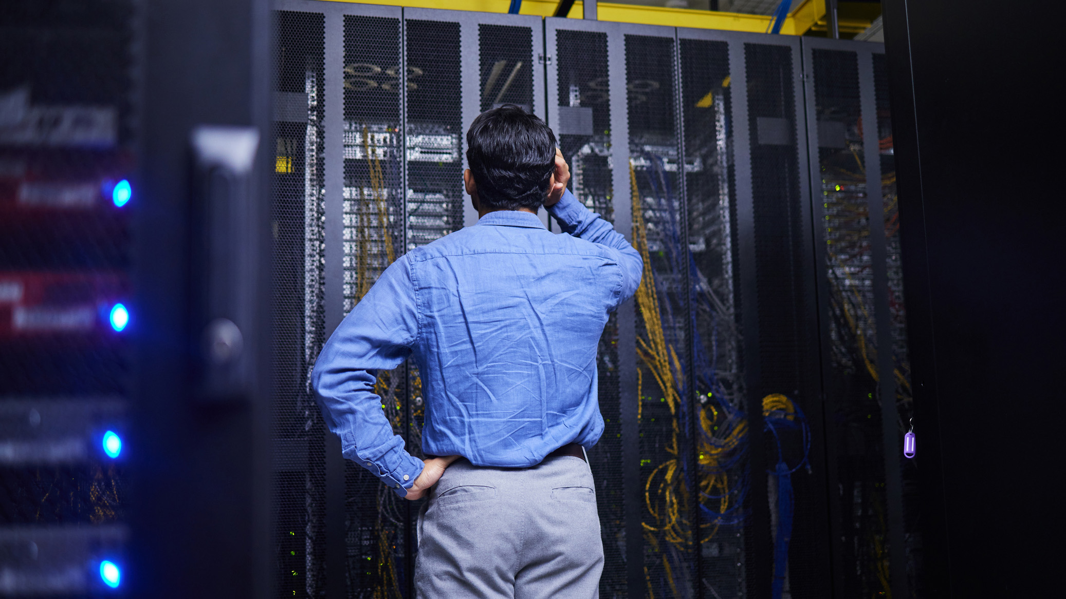 A businessman stood with his back to the camera facing a data center rack, with his arm to his head in frustration.