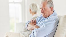 An older man sits on the sofa with his arms crossed, looking annoyed, while his wife sits in the background with her back to him.