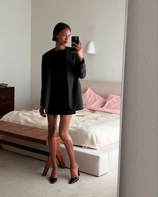 Woman wearing black mini dress and oversize black blazer taking mirror selfie.