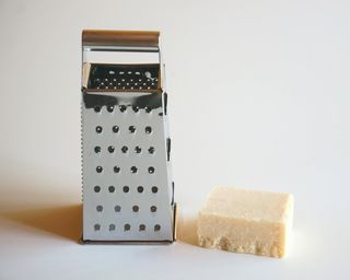 a metal box grater sat next to half a block of cheese on a white background