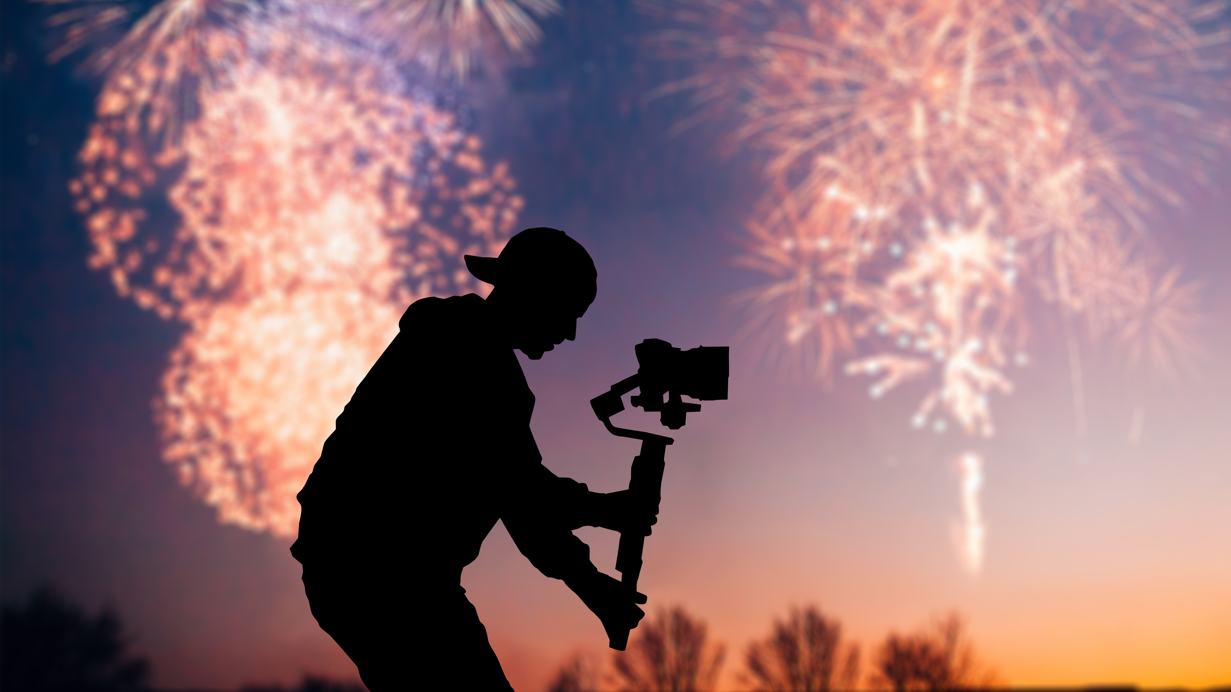 A videographer holds a camera on a gimbal with fireworks in the background
