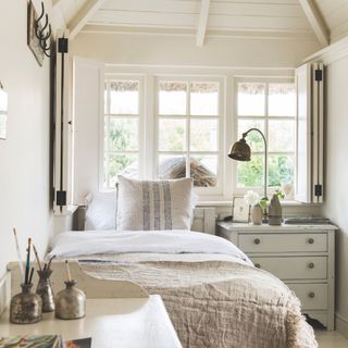 The bedroom has a neutral decor with a single bed and a chest of drawers with a lamp on top in front of the window.