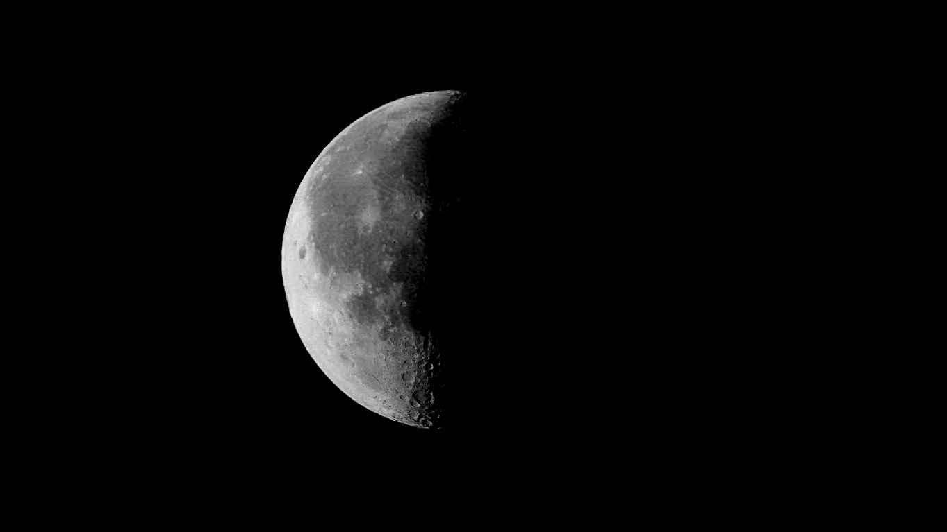 A picture of the moon with half its disk in shadow during its third quarter lunar phase. The lit half of the moon features darker regions known as Mare, or seas, along with craters, and is suspended against a black sky.