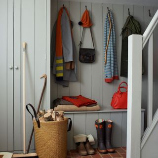 Neutral tongue and groove panelled wall hallway, stairs, brick terracotta brick floor, basket of firewood logs, coat hooks, storage bench, boots