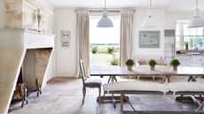 A large kitchen diner with a large dining table with cushioned wooden benches, flagstone floor and a huge stone fireplace feature to the left. Behind is a white AGA, and open doors with neutral curtains out to a large garden. Above are three large white lamps.