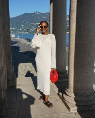 fashion influencer wanita Taffy Msipa tersenyum sambil berpose dengan pemandangan pegunungan yang indah di belakangnya mengenakan gaun putih lengan panjang tipis, tas merah, dan sandal Birkenstock hitam