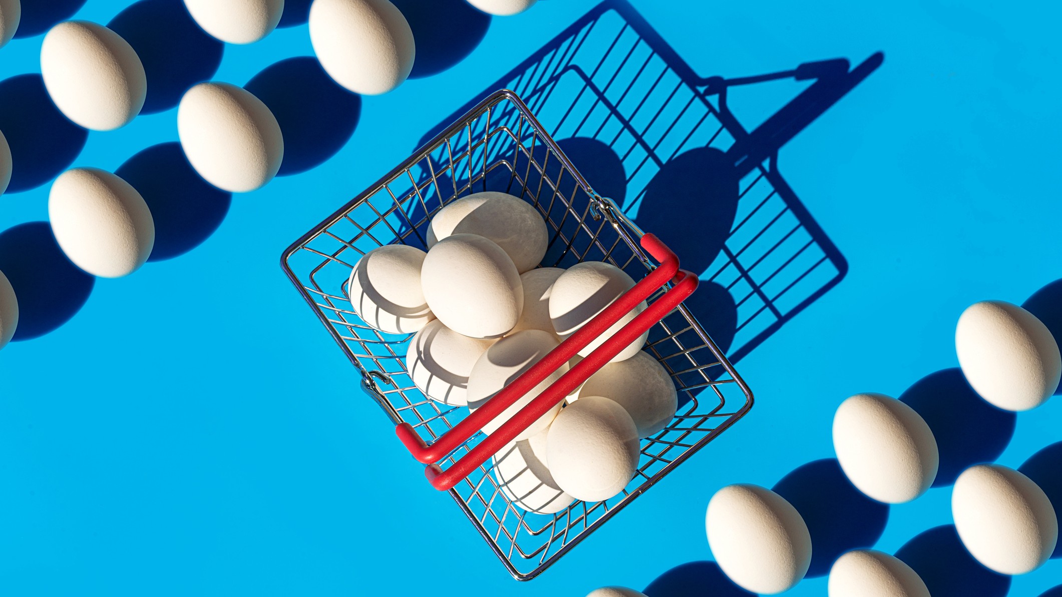 A wire basket full of eggs against a blue background. Other eggs are lined up in rows nearby. Conveys the concept of putting all of your eggs in one basket, or lack of diversification.