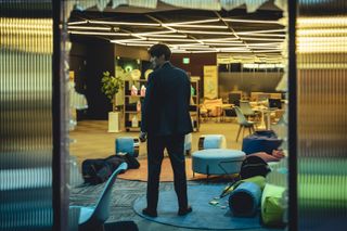 A man is seen through the broken frame of a glass wall, looking over his shoulder while standing in an office with toppled furniture around him, in 'Mercy for None.'