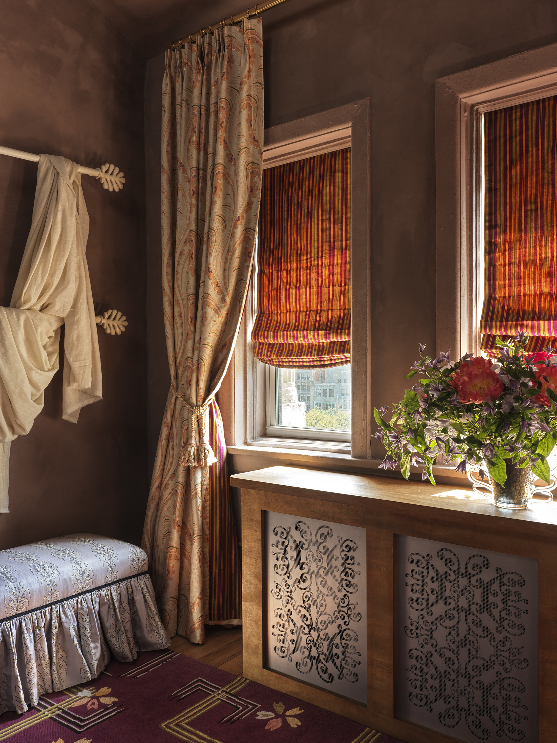 A view of a radiator below two windows, made with metal panels with a delicate pattern inspired by lace.