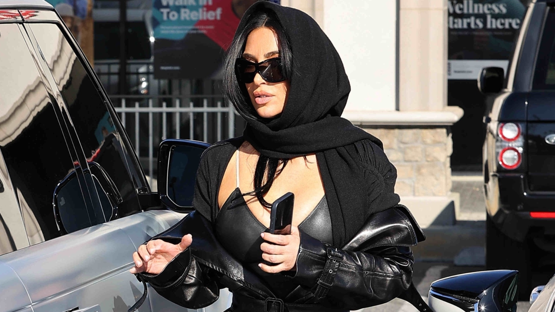Kim Kardashian goes undercover in all-black attire, including the leather trench coat trend and a peekaboo bra