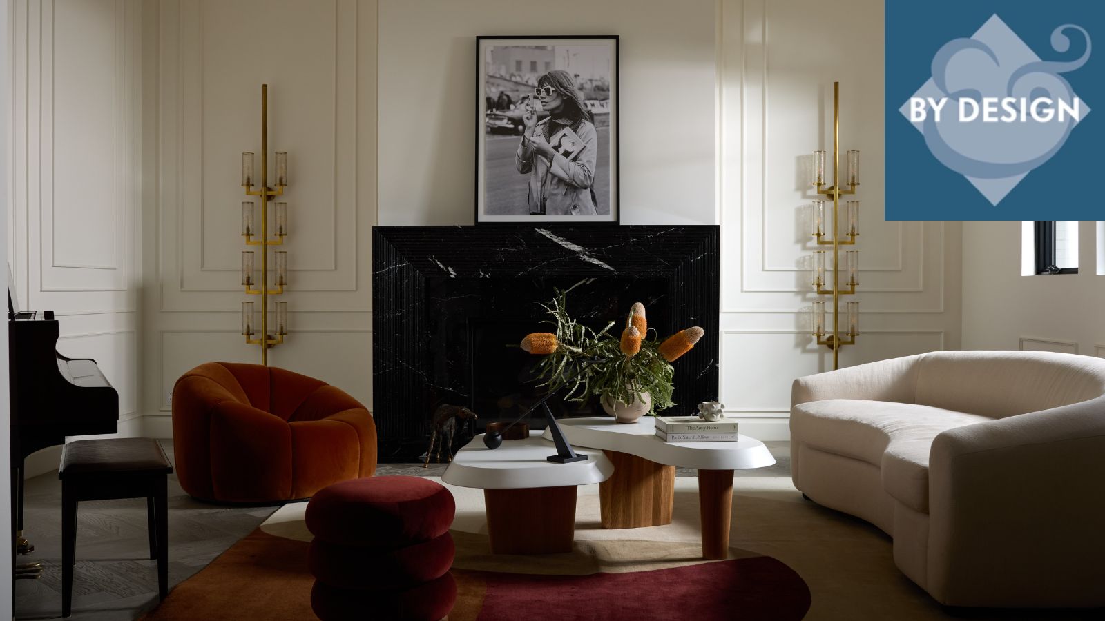 Modern living room with black marble fireplace, artwork on mantel, two sculptural floor lamps either side. Curved cream sofa, red lounge chair and ottoman, marble and wooden coffee table surround it