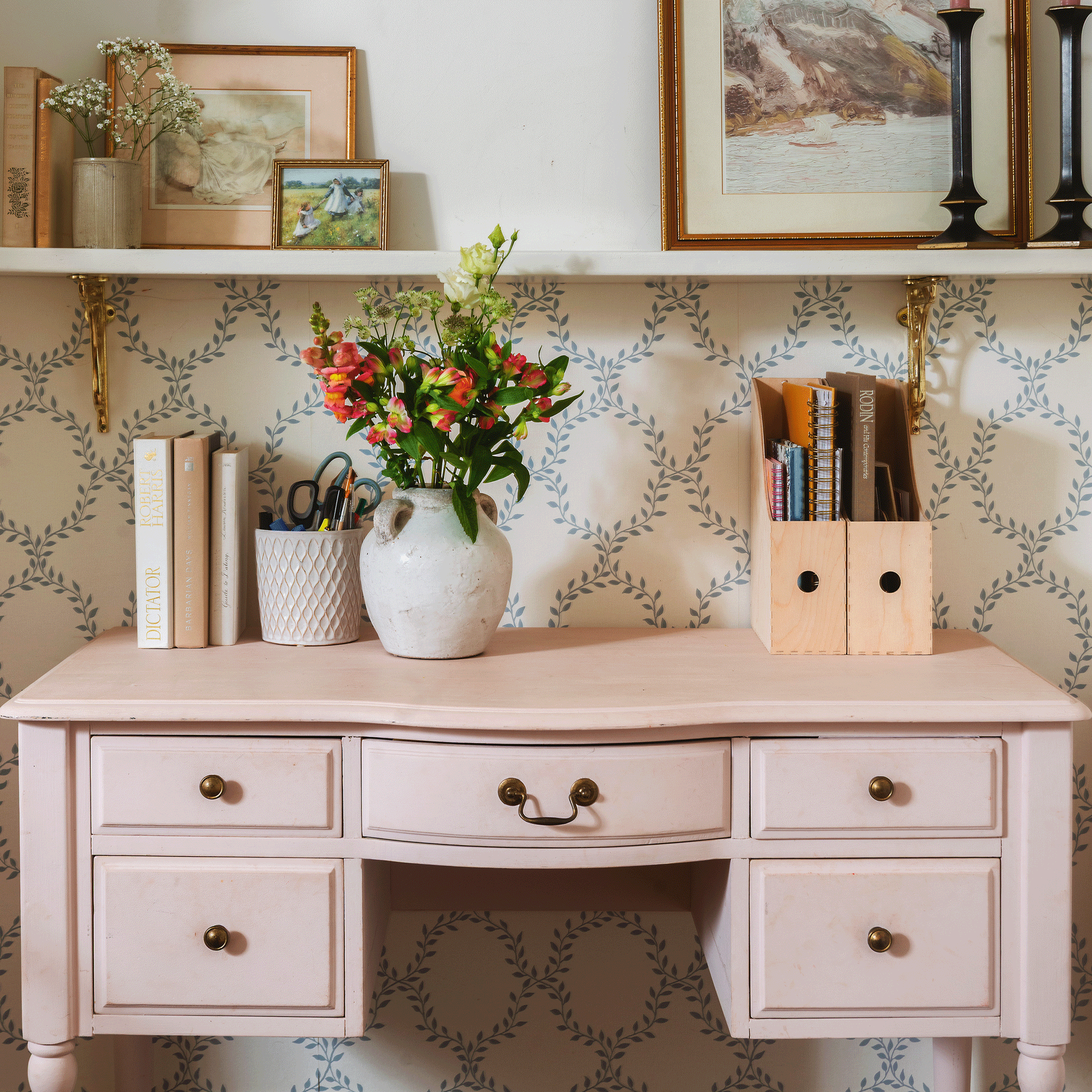 a pink desk with trellis wallpaper behind it with magazine racks and vase of flowers and a shelf above