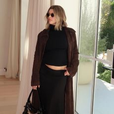 Sofia Richie Grainge poses indoors wearing a black turtleneck top with a black maxi skirt, a brown suede trench coat and a black bag with chain detailing.