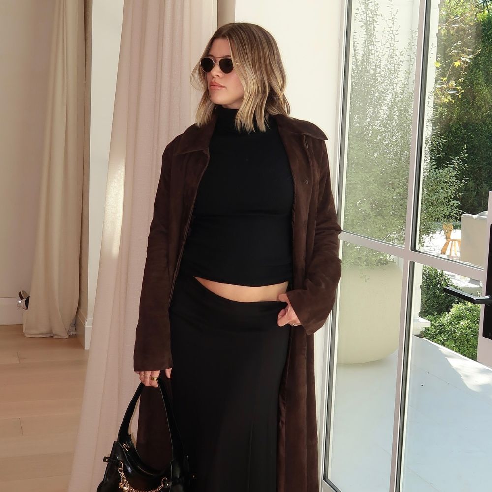 Sofia Richie Grainge poses indoors wearing a black turtleneck top with a black maxi skirt, a brown suede trench coat and a black bag with chain detailing.