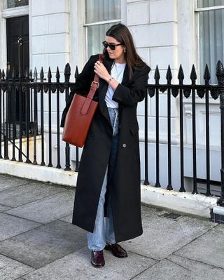 British influencer Anna Newton poses on a London sidewalk wearing black sunglasses, a long black coat, brown shoulder tote bag, white tee, straight-leg jeans, and brown loafers