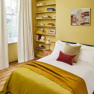 yellow bedroom with open shelving in alcove, white bedding with yellow blanket, coral stool, white desk lamp, wooden floor boards, white curtains, art work
