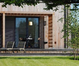 The corner of a timber-clad new build, with a timber veranda providing shelter and shading around patio doors and a patio area. A newly lawned garden in front features a further patio with timber loungers. The timber cladding was supplied by Vastern Timber.