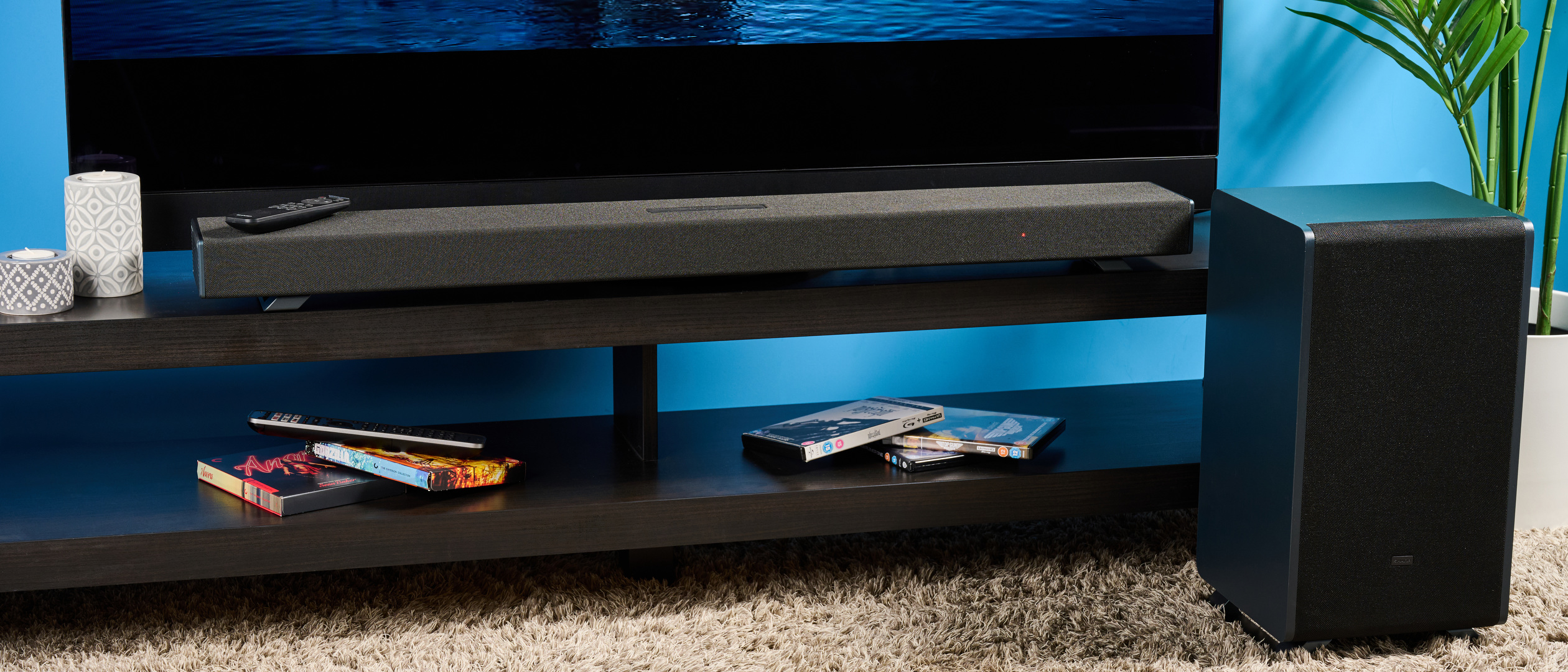 the Sharp 3.1.2 HT-SBW53121 Soundbar in black with a remote, subwoofer, against a blue background next to a panasonic TV