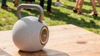 A grey Lenco Orbita-360 speaker on a table outside with people visible standing on grass in the background.