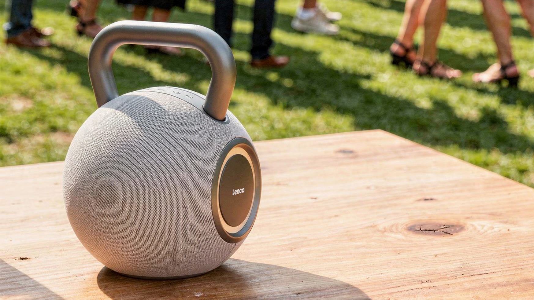 A grey Lenco Orbita-360 speaker on a table outside with people visible standing on grass in the background.