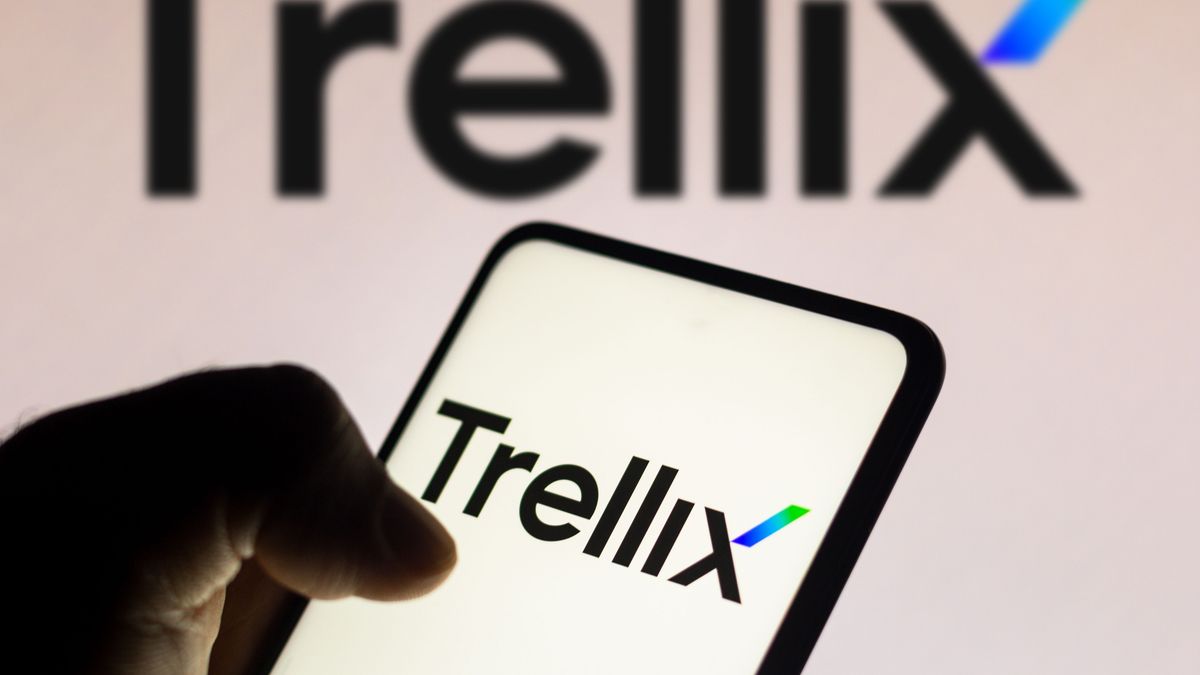 Trellix logo on a smartphone. Trellix is also written on a white wall behind the phone which is being held by a hand