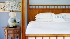 Blue bedroom with warm wood four-poster bed, white bedding, and circular patterned nightstand with a white lamp with blue and white patterned lampshade and blue hydrangeas. 