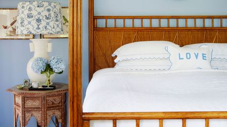 Blue bedroom with warm wood four-poster bed, white bedding, and circular patterned nightstand with a white lamp with blue and white patterned lampshade and blue hydrangeas. 