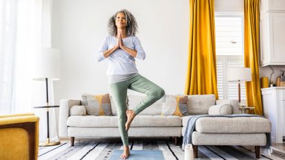 woman holding a tree yoga pose in a living room setting with a grey L-shape sofa behind her and a window with long yellow curtains.