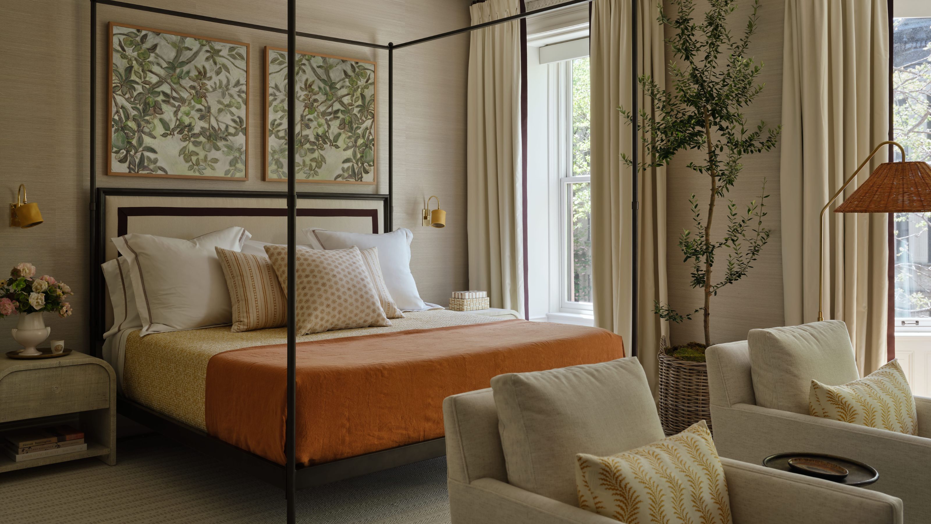 a large neutral primary bedroom with a four poster bed, green artwork, orange bed throw, a tall indoor plant and a seating area