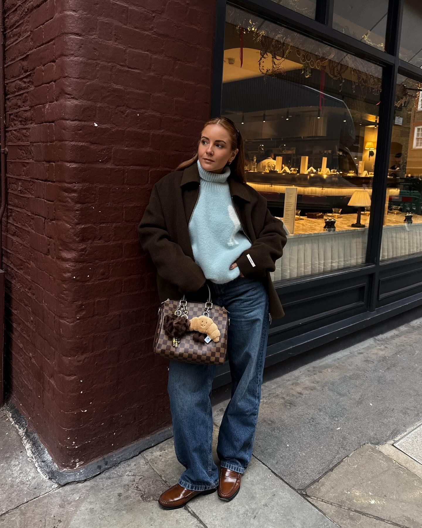 Fashion person Rebecca Ferraz Wyatt wears a brown loafer, blue denim jean, pale blue jumper and brown coat. She is wearing a brown coat and holding a brown LV speedy bag. She is stood leant on a red brick wall looking into the difference in a recent image from her Instagram.