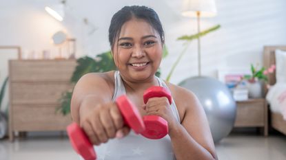 Smiling woman in domestic setting holding light dumbbells and punching towards the viewer 