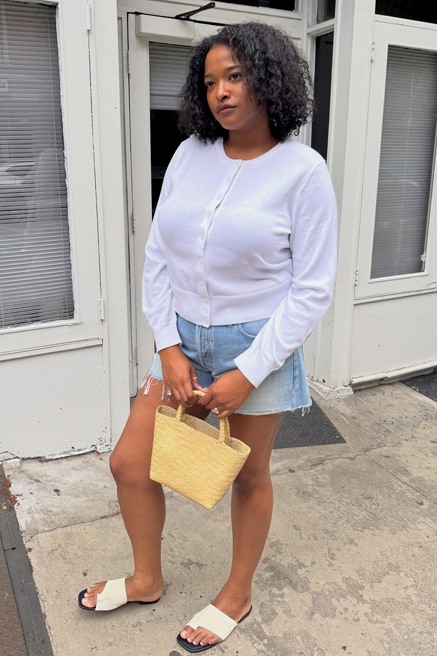 A woman wearing a white cardigan, denim shorts, white slide-on sandals, and a woven handbag.