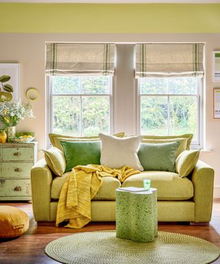 A green living area with lime green and beige walls, two windows with folded blinds, a lime green couch with green and beige throw pillows and a mustard throw draped on it and a green abstract coffee table